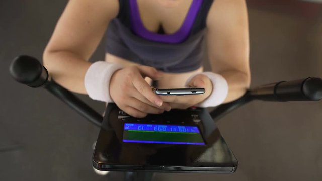 Plump female riding stationary bike and viewing photos on smartphone, fitness
