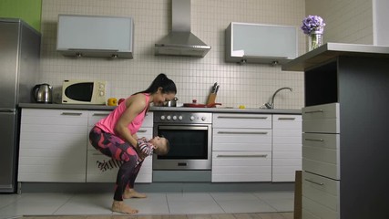 Happy fit mother doing squats lifting her baby boy at home in the kitchen. Sporty gorgeous female housewife making exercises together with her laughing infant child. Active family workout. Dolly shot. - Powered by Adobe