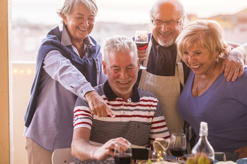 4 senior with white hair on the terrace at the table set laughing looking at the cell phone. Everyone in a group, together with a glass of wine, embrace each other and indicate the telephone.