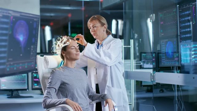 Woman Wearing Brainwave Scanning Headset Sits in a Chair while Scientist Adjusts the Device, Looks at Displays. In the Modern Brain Study Laboratory Monitors Show EEG Reading and Brain Model. 