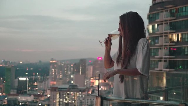 Happy woman drinking a glass of white wine while sitting in rooft bar in the city or on the terrace with cityscape at sunset. Vacation,happiness,freedom concept.