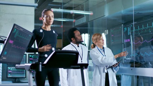 Beautiful Woman Athlete Runs on a Treadmill with Electrodes Attached to Her Body, while Two Scientists Supervise Watching EKG Data Showing on Laboratory Monitors.
