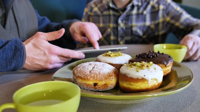 Electronics Technology Concept Men Viewing Discussing Media on Tablet Computer Screen During Casual Meeting WIth Paczki Cream Jelly Filled Donut Pastry Plate Hot Tea Cups