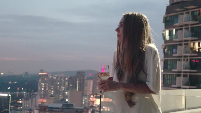 Happy woman drinking a glass of white wine while sitting in rooft bar in the city or on the terrace with cityscape at sunset. Vacation,happiness,freedom concept.