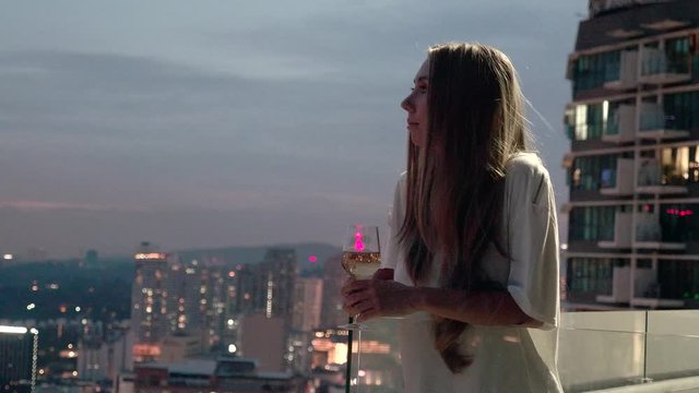 Happy woman drinking a glass of white wine while sitting in rooft bar in the city or on the terrace with cityscape at sunset. Vacation,happiness,freedom concept.