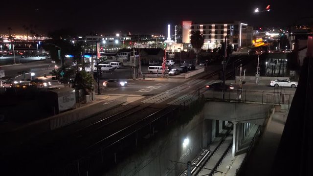 cars go through a railroad crossing and over a subway trench, in the background a plane is landing over the buildings. This is night, we are in  a big city. 