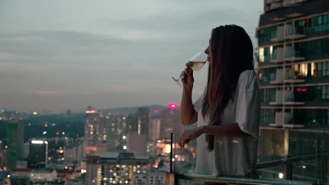 Happy woman drinking a glass of white wine while sitting in rooft bar in the city or on the terrace with cityscape at sunset. Vacation,happiness,freedom concept.