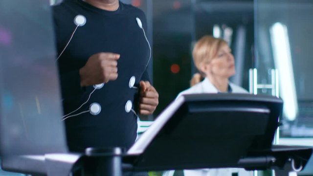 Male Athlete Runs on a Treadmill with Electrodes Attached to His Body while Sport Scientist Supervises Reads EKG Status. In the Background Laboratory with High-Tech Equipment.