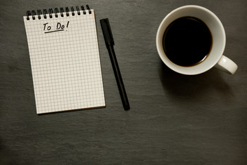 To do list on gridded spiral notepad with cup of coffee, on slate table top - from above, minimal