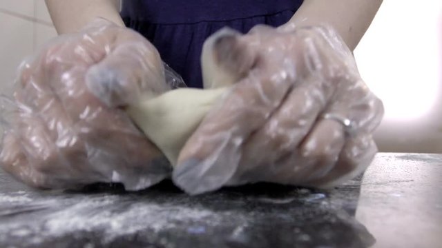 Dough is being kneaded by young female baker on bright kitchen, close up. Woman is using both of her hands in white gloves, whith beautiful manicure, to beat the pastry and to prepare it for baking on