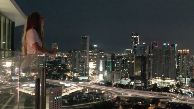 Happy woman drinking a glass of white wine while sitting in rooft bar in the city or on the terrace with cityscape at sunset. Vacation,happiness,freedom concept.