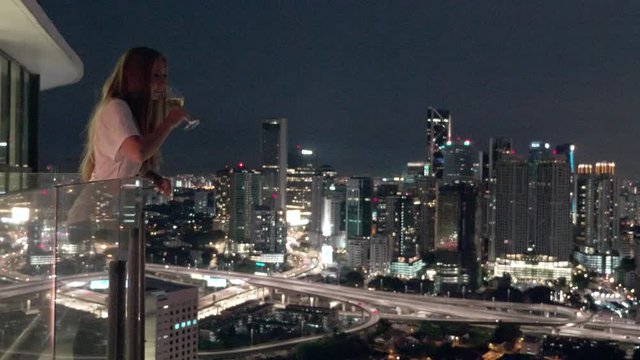 Happy woman drinking a glass of white wine while sitting in rooft bar in the city or on the terrace with cityscape at sunset. Vacation,happiness,freedom concept.