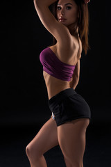 Fototapeta na wymiar Young beautiful dark-haired woman in dark purple sports bra and black short shorts seductively posing with her back against a black background