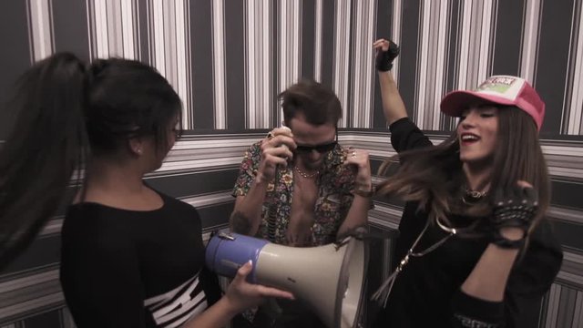 Rapper caucasian tattooed guy in sunglasses with sexy girls in black dresses dancing and rapping into megaphone at apartment, striped wallpaper