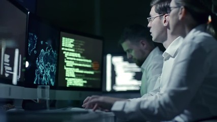 Professional team of cyber security specialists writing program code and working with holographic map on computers in IT office at night