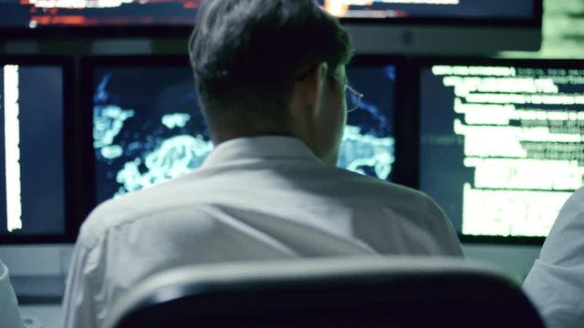 Rear view of professional team of IT security specialists talking in front of multi-display workstation in dark office at night; computer code and holographic world map on display