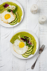 Spinach green crepes (pancakes) with fried egg, avocado and leaves of mix of salad on ceramic plate on white wooden background. Сoncept of healthy breakfast. Selective focus. Top view. Copt space.