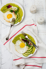 Spinach green crepes (pancakes) with fried egg, avocado and leaves of mix of salad on ceramic plate on white wooden background. Сoncept of healthy breakfast. Selective focus. Top view. Copt space.