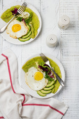 Spinach green crepes (pancakes) with fried egg, avocado and leaves of mix of salad on ceramic plate on white wooden background. Сoncept of healthy breakfast. Selective focus. Top view. Copt space.