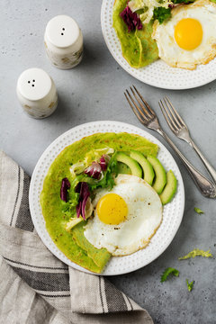 Spinach green crepes (pancakes) with fried egg, avocado and leaves of mix of salad on ceramic plate on gray concrete background. Сoncept of healthy breakfast. Selective focus. Top view. Copt space.