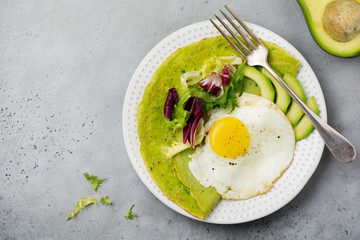 Spinach green crepes (pancakes) with fried egg, avocado and leaves of mix of salad on ceramic plate on gray concrete background. Сoncept of healthy breakfast. Selective focus. Top view. Copt space.