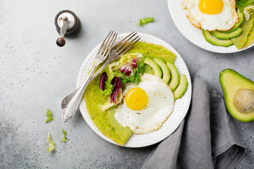 Spinach green crepes (pancakes) with fried egg, avocado and leaves of mix of salad on ceramic plate on gray concrete background. Сoncept of healthy breakfast. Selective focus. Top view. Copt space.