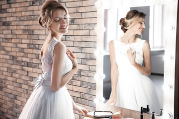 Naklejka premium Beautiful young bride in white wedding dress with makeup created by professional artist near mirror indoors