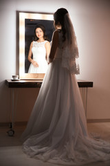 Fototapeta premium Beautiful young bride in white wedding dress with makeup created by professional artist near mirror indoors