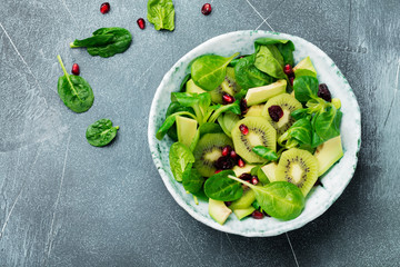 Salad of spinach baby leaves, watercress, kiwi, avocado and pomegranate in old ceramic plate on gray concrete background. Selective focus. Top view. Copy space.