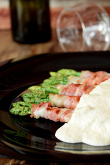 Green asparagus wrapped into smoked bacon covered with white sauce on a black plate