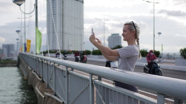 Slim woman with a pigtail blonde in a gray T-shirt standing on the bridge and take a pictures by smartphone over background sea and city.