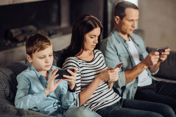 Modern realities. Pleasant young family sitting on the couch and using their phones, being addicted to them, while noticing nothing going on around