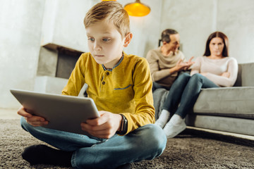 Being lonely. Sad little boy sitting cross-legged on the floor and playing on his tablet while his parents sitting on the couch and quarrelling
