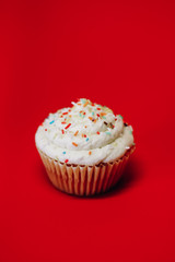 Traditional American cupcake with vanilla cream cheese frosting with colorful sprinkles on solid red background