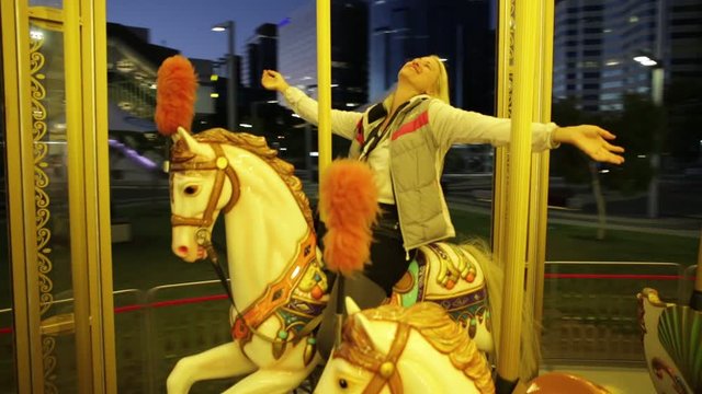 Happy blonde caucasian woman riding on white horse of Perth city carousel in Western Australia at night. Leisure and recreational concept.