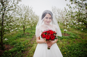 Portrait of a gentle young bride holding red bouquet in the blossoming garden on her wedding day.