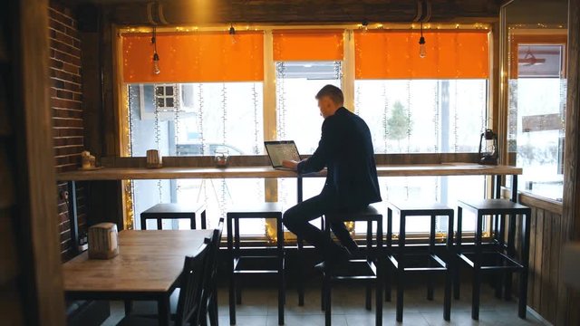 Working from cafe. Cheerful young man in smart casual wear looking at laptop and sitting near window in creative office or cafe