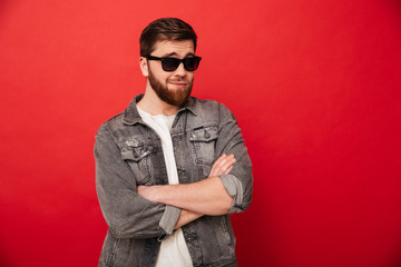 Obraz premium Portrait of irresistible young guy wearing sunglasses and denim smiling and posing with hands crossed, isolated over red background