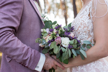Wedding. The girl in a beige dress and a guy in a suit are holding a beautiful bouquet of white, purple, lilac flowers and greenery. Wedding rustic bouquet with lilac roses. Wedding details