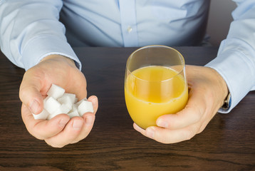 Man sits at the table and has a glass of orange juice in front of him in his hand he holds many pieces of sugar cubes