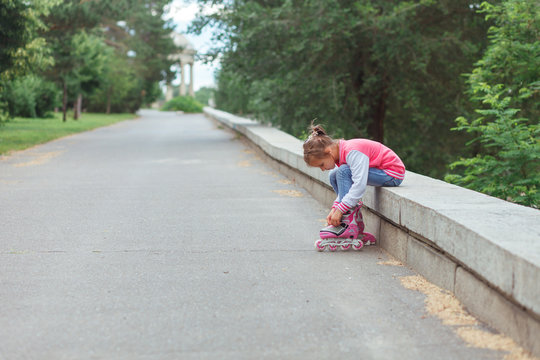 Little girl sitting on parapet and putting on rollers in the park