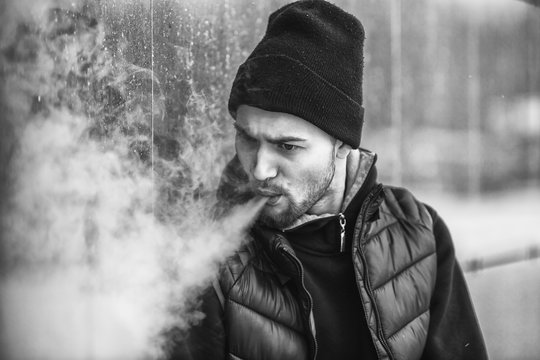 Vape man. Portrait of a handsome young white guy in black waistcoat and modern cap vaping an electronic cigarette opposite the futuristic urban background. Black and white.