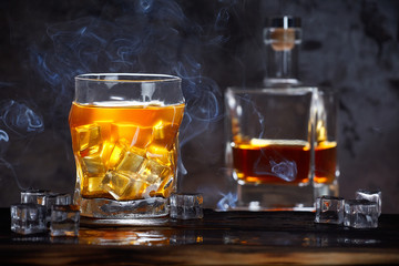 a glass of whiskey with ice in smoke. Ice on wooden background. A bottle of whiskey in the background