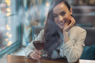 Wine affect. Gorgeous young positive woman touching glass of wine while putting head on hand and posing at table