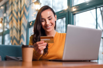 Payment method. Merry enthusiastic nice woman reading credit card number while staring down and working on laptop