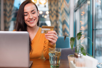 Credit card number. Joyful jolly beautiful woman examining credit card number while staring at it and laptop lying on table