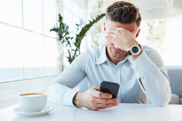 Photo of adult upset man 30s sitting alone in city cafe with cup of cappuccino, and propping up his...