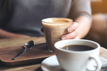 Closeup image of Americano coffee cup and woman's hand holding latte coffee cup on vintage wooden table in cafe