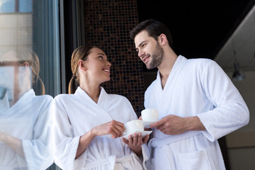 Obraz premium couple in bathrobes holding cups of tea and smiling each other in spa center