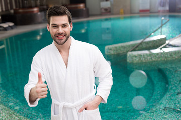 handsome young man in bathrobe showing thumb up and smiling at camera in spa center